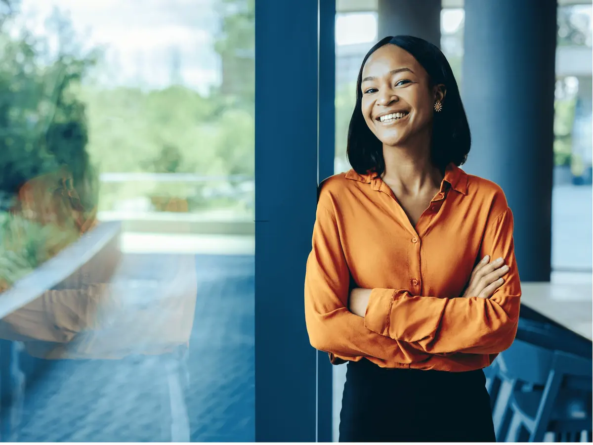A smiling professional stands with her arms folded in a sunlit office lobby